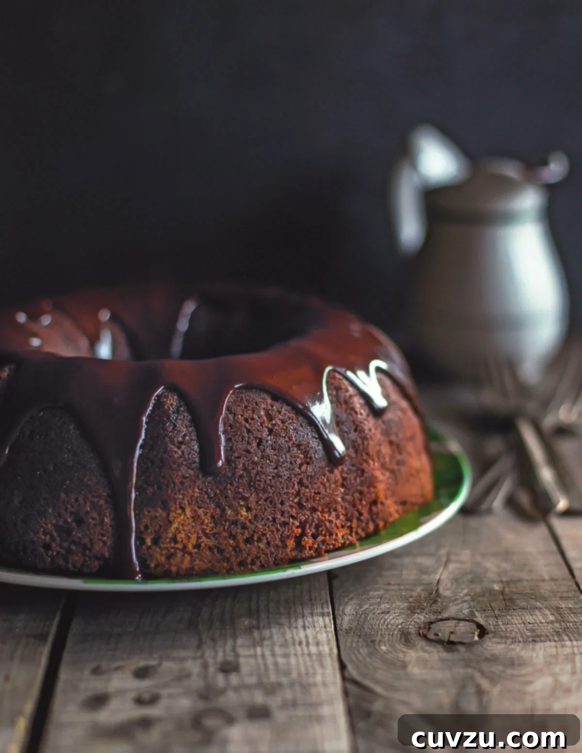 Decadent Chocolate Zucchini Cake 2 A wooden table with a pitcher and a green plate holding a chocolate bundt cake drizzled in chocolate ganache. This luxurious cake is the epitome of summer indulgence, combining rich chocolate with the hidden goodness of zucchini for an irresistibly moist texture.
