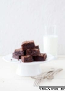 A white cake stand of a pile of one-pot cocoa brownies, a glass of milk, on a white table