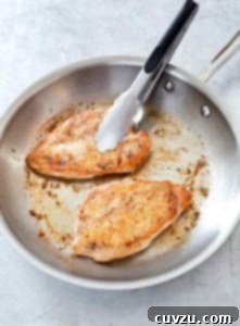 Two pan seared chicken breasts in a stainless steel skillet with a pair of tongs resting on one of the chicken breasts.