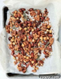A layer of assorted candied spiced nuts and rosemary sprigs on a sheet of parchment paper resting in a sheet pan, perfectly prepared.