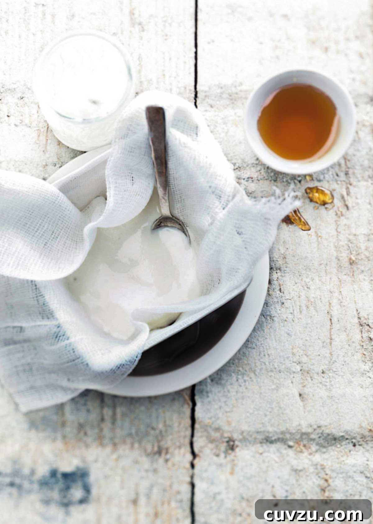 DIY Greek Yogurt Made Easy 2 Homemade Greek yogurt being strained through cheesecloth into a white bowl with a spoon and a small bowl of honey beside it.