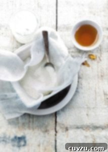 Homemade Greek yogurt being strained through cheesecloth into a white bowl with a spoon and a small bowl of honey beside it.