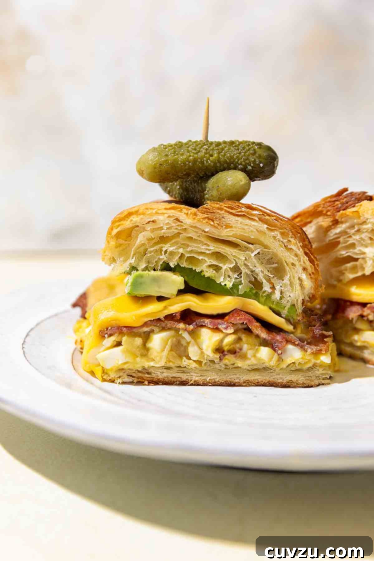 Egg Salad Reinvented 8 A halved croissant sandwich with egg salad, bacon, cheese, avocado, and small pickles on a white plate.
