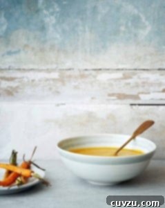 Gwyneth's Golden Carrot Ginger Soup 3 A white bowl filled with creamy carrot ginger soup, with a spoon resting inside and a plate of carrots beside it.