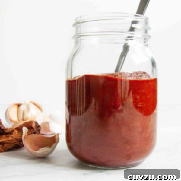 A jar of homemade adobo sauce with a spoon resting inside and garlic cloves and dried chipotles on the side.