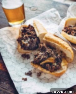 Two savory Philly cheesesteak sandwiches halved on a foil wrapper, next to a glass of beer, highlighting their deliciousness.