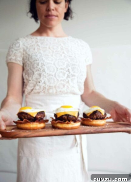 A woman holding a wooden tray with 3 breakfast burgers topped with eggs.