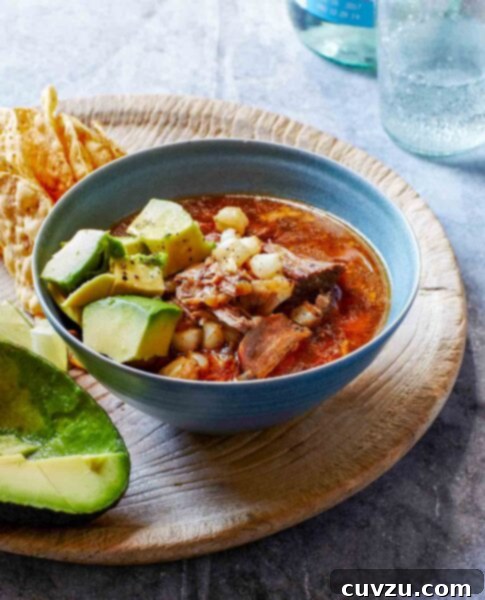 A bowl of slow cooker pork posole with tender pork, hominy, and a rich red broth, garnished with fresh avocado and served with tortilla chips.