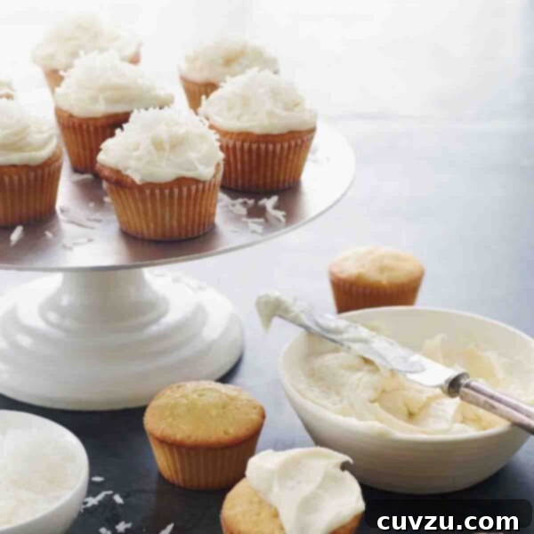 Cake stand with 7 frosted coconut cupcakes, four unfrosted cupcakes on the table, and a bowl of frosting.