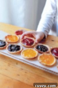 A tray of mini jam tarts with various fillings like orange, strawberry, and blueberry, with a child's hand reaching for one.