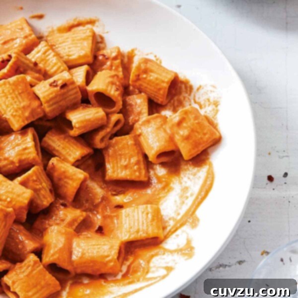 A white serving bowl and smaller white bowl partially filled with rigatoni with vegan vodka sauce and a glass of water on the side.