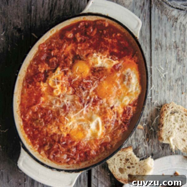 An oval casserole dish of acquacotta - eggs poached in a tomato and onion sauce -- with a piece of bread, plate, and utensils on the side.