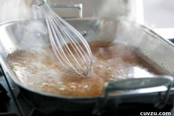A person whisking gravy with a large metal balloon whisk in a metal roasting pan.