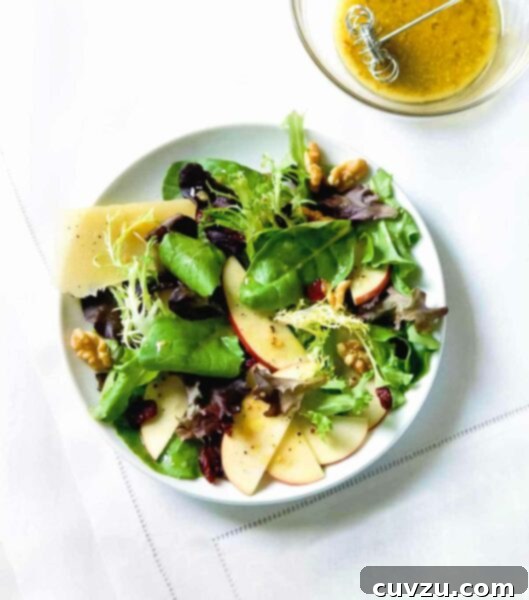 A large white bowl brimming with an artistic arrangement of apples, Parmesan, walnuts, and mixed green salad, accompanied by a small, inviting bowl of honey dressing.