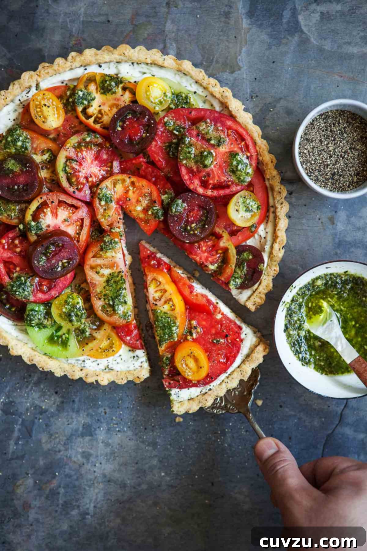 Savory Garden Tomato Galette 2 A person carefully serving a slice of fresh tomato tart from a pan, with a small bowl of pesto vinaigrette and black pepper shaker on the side, highlighting the tart's golden crust and colorful tomato slices.