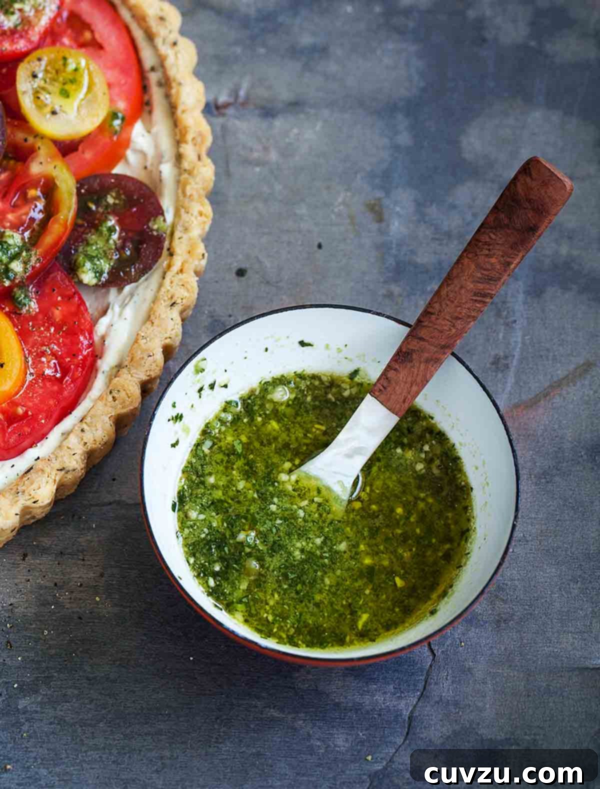 Savory Garden Tomato Galette 4 A small glass bowl filled with vibrant green pesto vinaigrette, with a small spoon resting inside, placed next to a slice of fresh tomato tart on a plate.