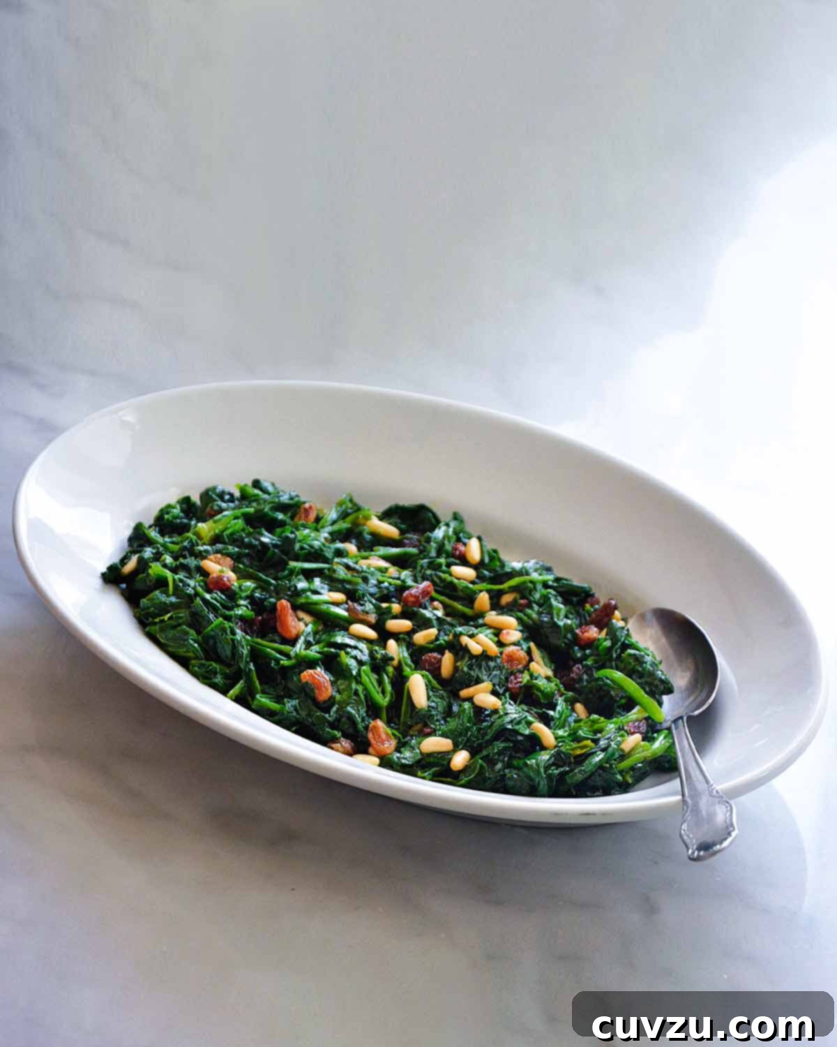A white oval dish filled with cooked spinach with raisins and pine nuts sprinkled over the top and a spoon resting in the dish.