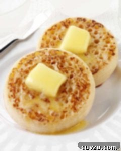 Two golden-brown English crumpets, each generously topped with a melting pat of butter, resting on a pristine white plate.