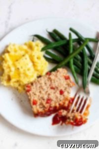 A slice of turkey meatloaf on a white plate with mashed potatoes, green beans, and fork resting on the side.