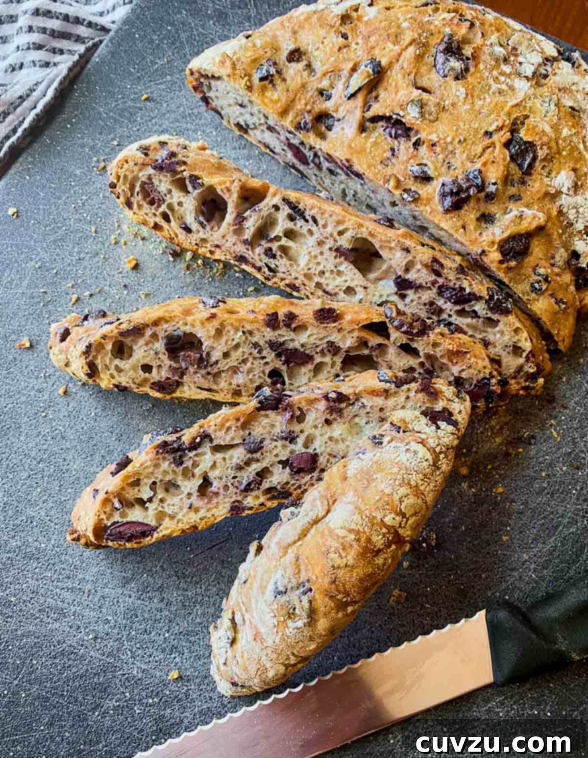 Easy Mediterranean Olive Loaf 3 A beautifully rustic Jim Lahey no-knead olive bread, cut into four generous slices, showcasing its invitingly airy crumb and abundance of black Kalamata olives. It is presented on a vibrant blue cutting board, accompanied by a folded napkin, a specialized bread knife, and a small mound of sliced black olives, highlighting the bread's rich flavor profile.