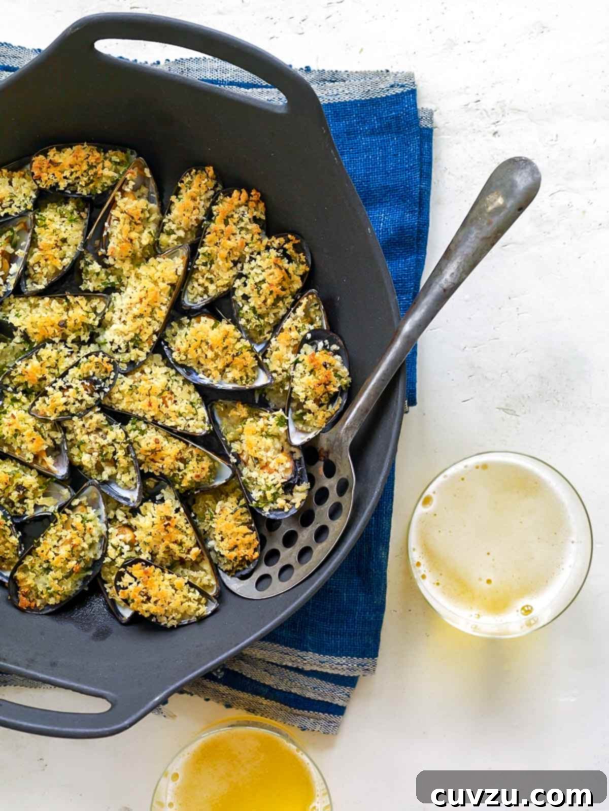 Crispy Breadcrumb Mussels 2 Golden-brown baked mussels topped with crisped breadcrumbs, served in a cast-iron pan with a serving spoon on a vibrant blue tablecloth.