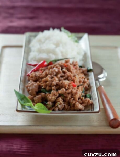 A wooden cutting board with a rectangular plate and spoon on top, the plate has a mound of rice on the back half. On the front half, there's a pile of ground pork, basil, and peppers.