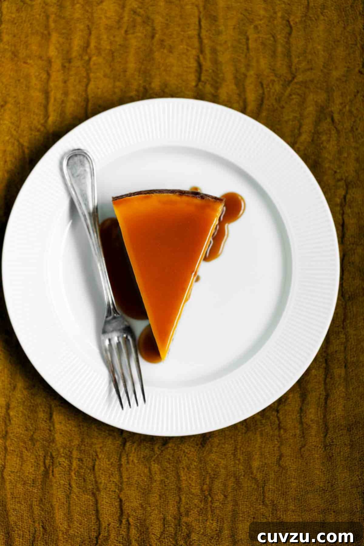 Velvet Pumpkin Cheesecake 4 A slice of pure pumpkin cheesecake on a white plate, drizzled with caramel glaze and accompanied by a fork, ready to be enjoyed.