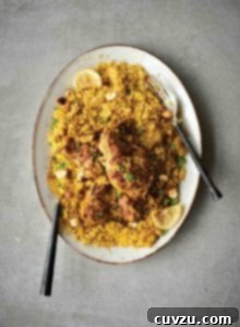 A white oval platter filled with couscous that is topped with Middle Eastern chicken thighs, lemon slices, and chopped parsley.