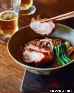 A bowl of duck soba with scallions on a wooden surface with a piece of duck being lifted out with chopsticks.