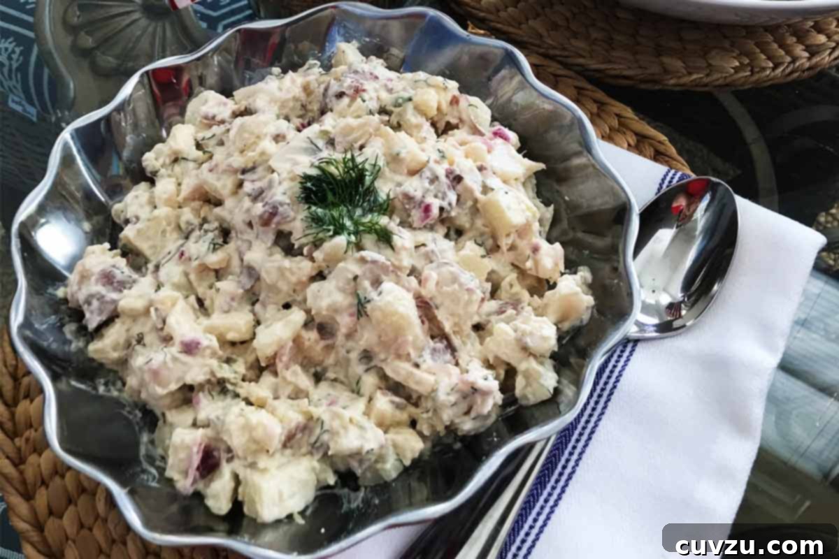 Crisp Apple Potato Salad 5 A silver bowl filled with potato salad with apples topped with dill and a spoon and striped napkin on the side.