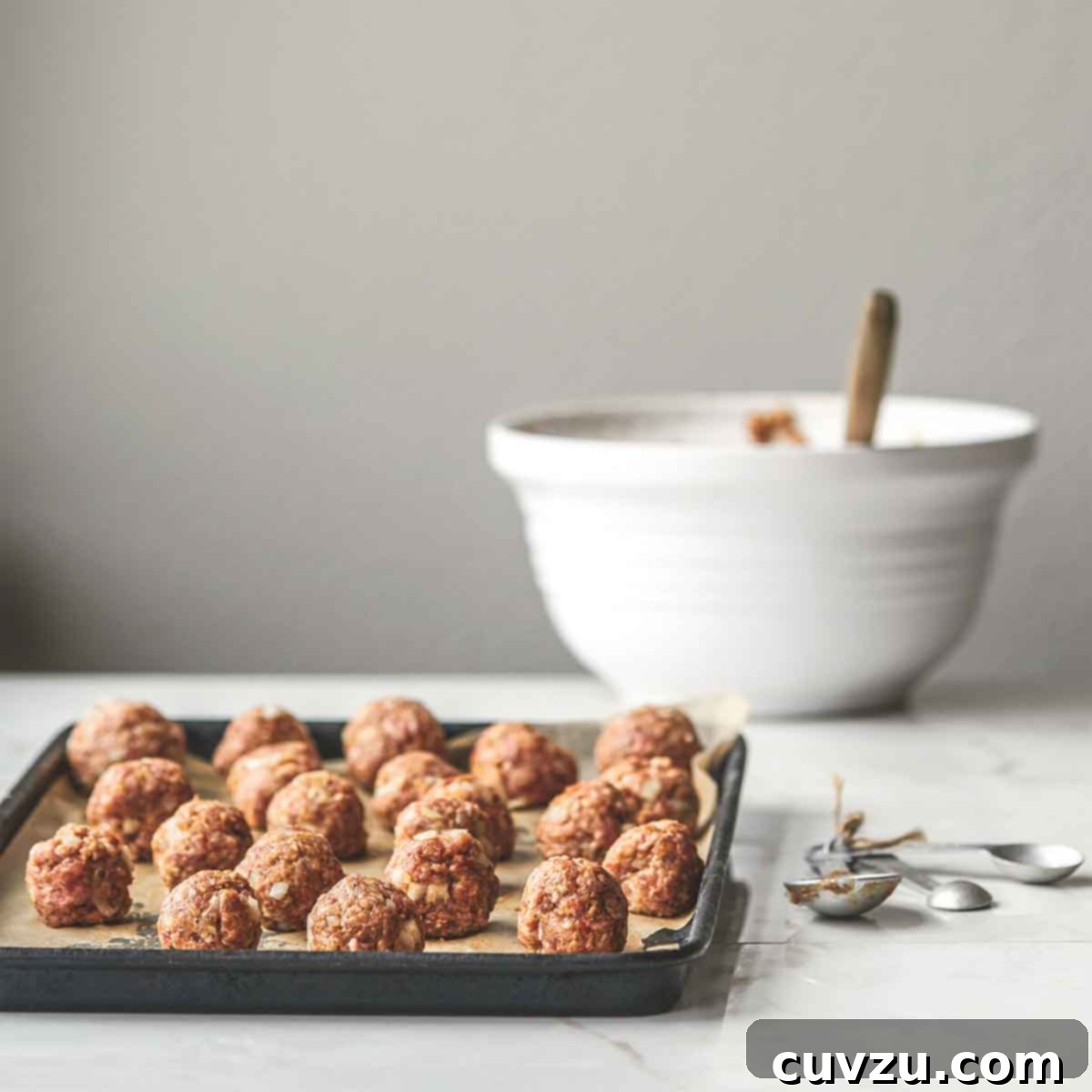 A tray of unbaked bbq meatballs with a set of measuring spoons beside it and a bowl of barbecue sauce in the background.
