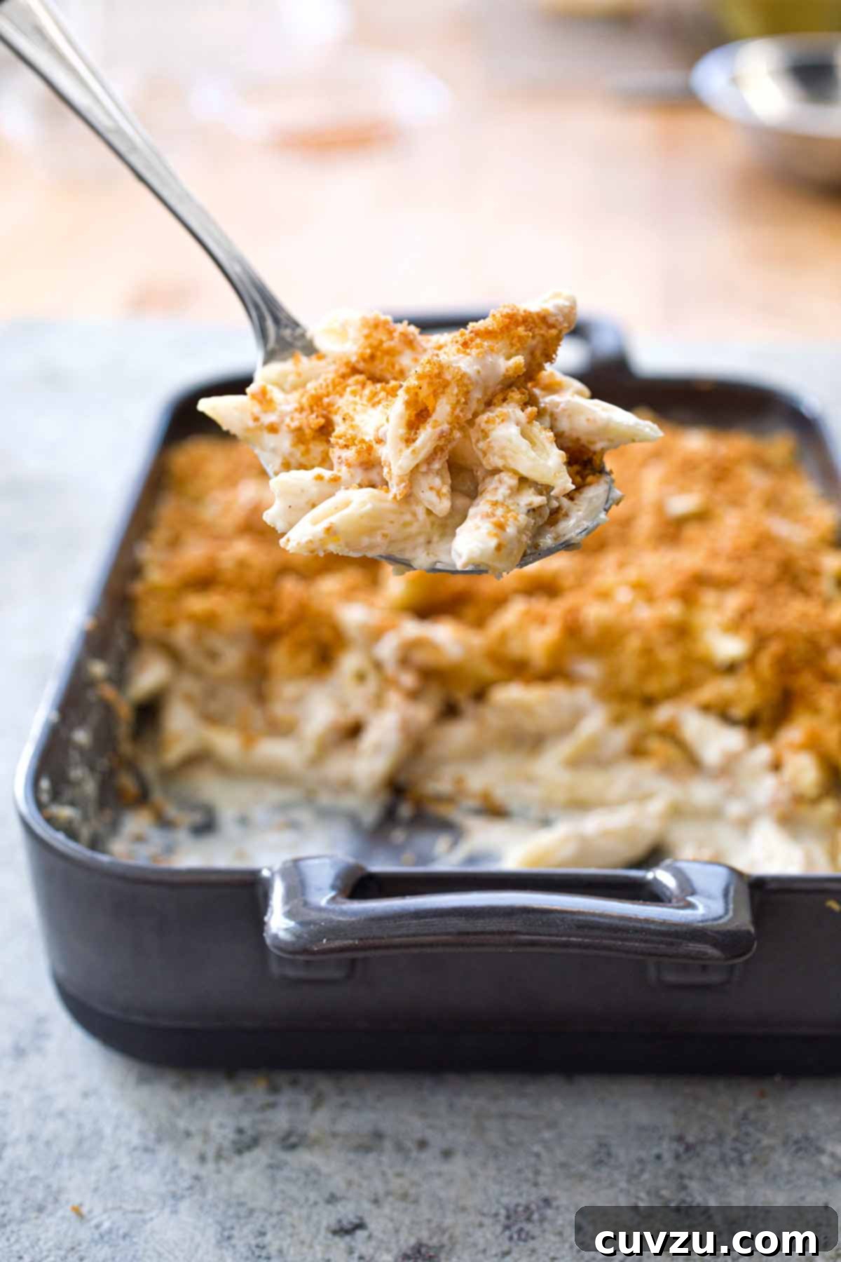 A dark grey rectangular baking dish half filled with four cheese macaroni and cheese and a metal spoon lifting a scoop out.