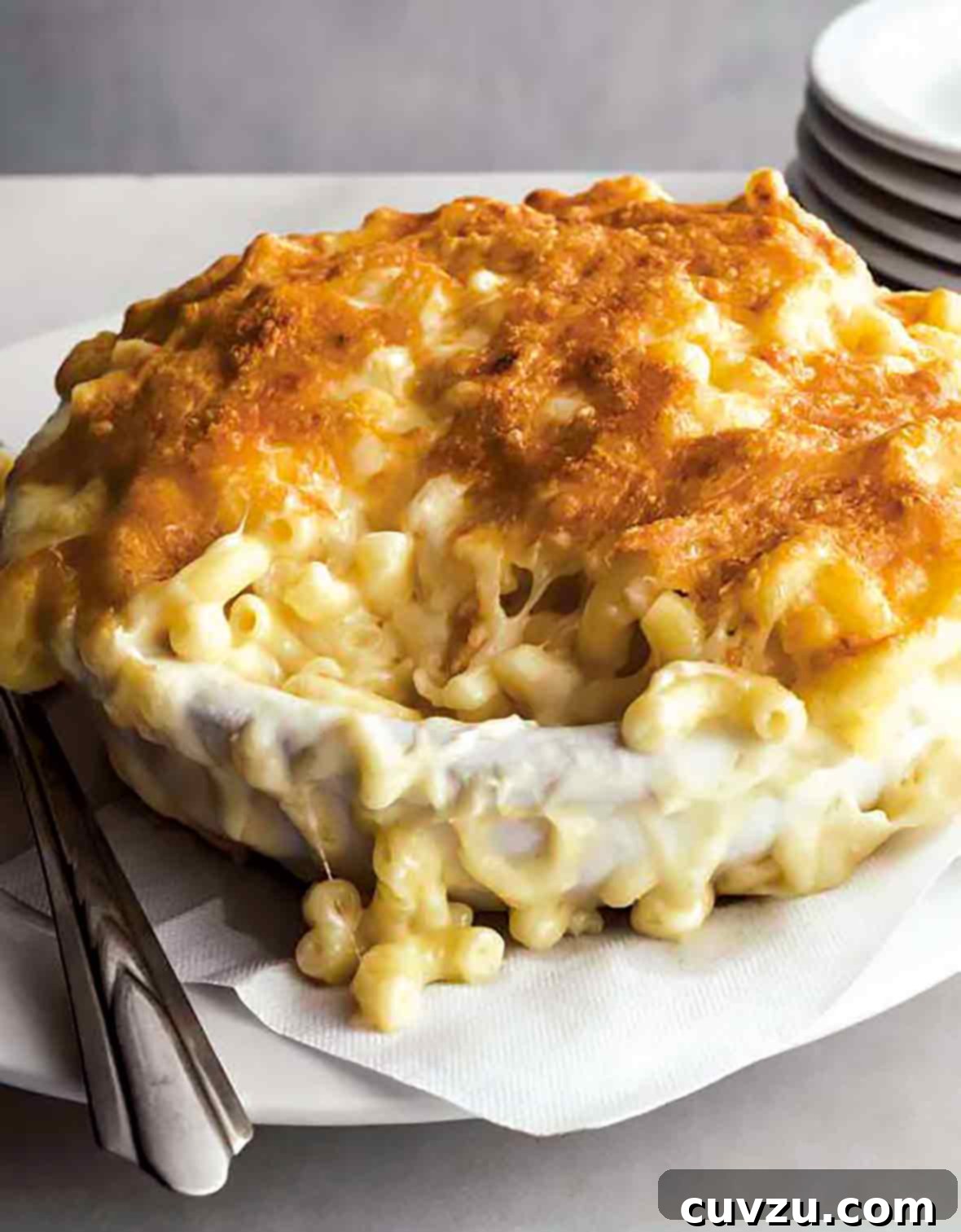 A dish of macaroni au gratin on a white paper napkin on a white plate with a stack of plates beside it.