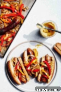 A serving of sheet pan sausage, peppers, and onions on a white plate, with a jar of mustard visible beside it.