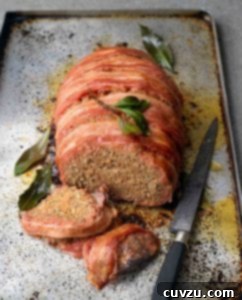 A partially sliced bacon-wrapped meatloaf on a rimmed baking sheet with a knife beside it and bay leaves scattered around it.