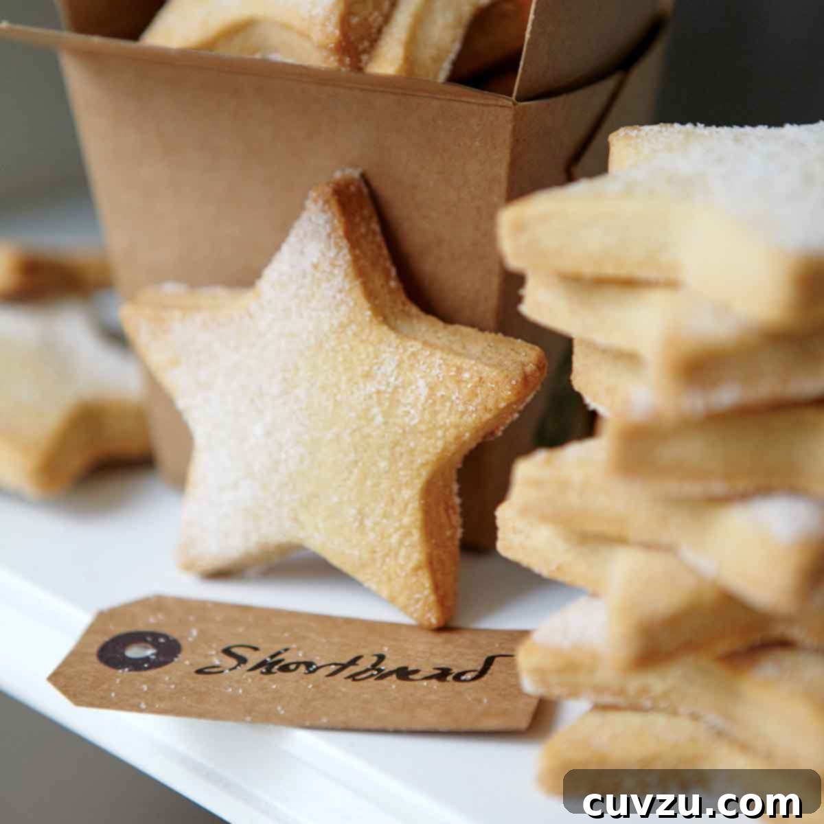 A stack of golden, star-shaped classic shortbread cookies, lightly dusted with sugar, next to a brown takeout container and a gift tag.