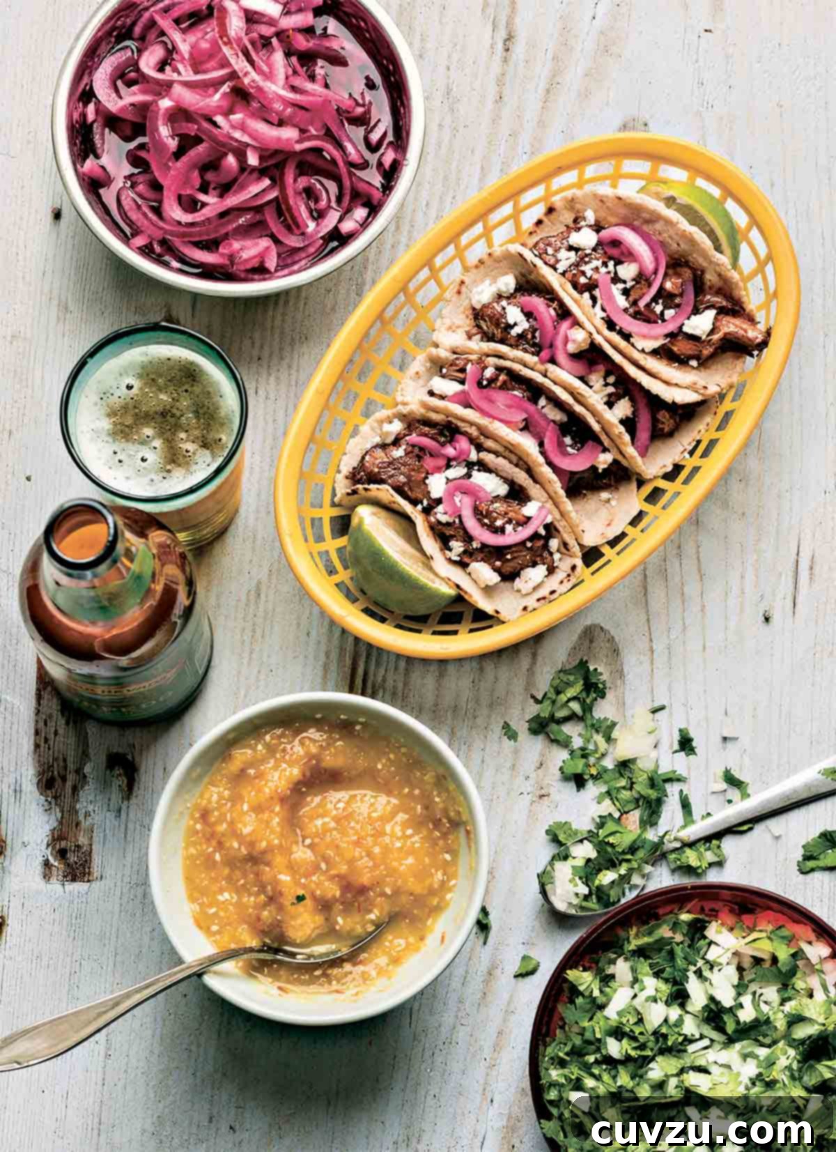 Four beef chili tacos and two lime halves in a basket, with bowls of pickled onions, slaw, and a beer on the side.