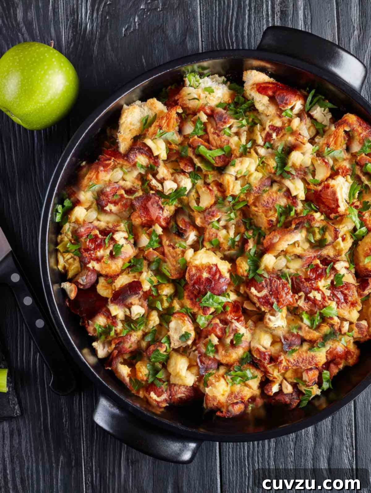 A black bowl of bread stuffing with herbs and bacon, alongside is a green apple.