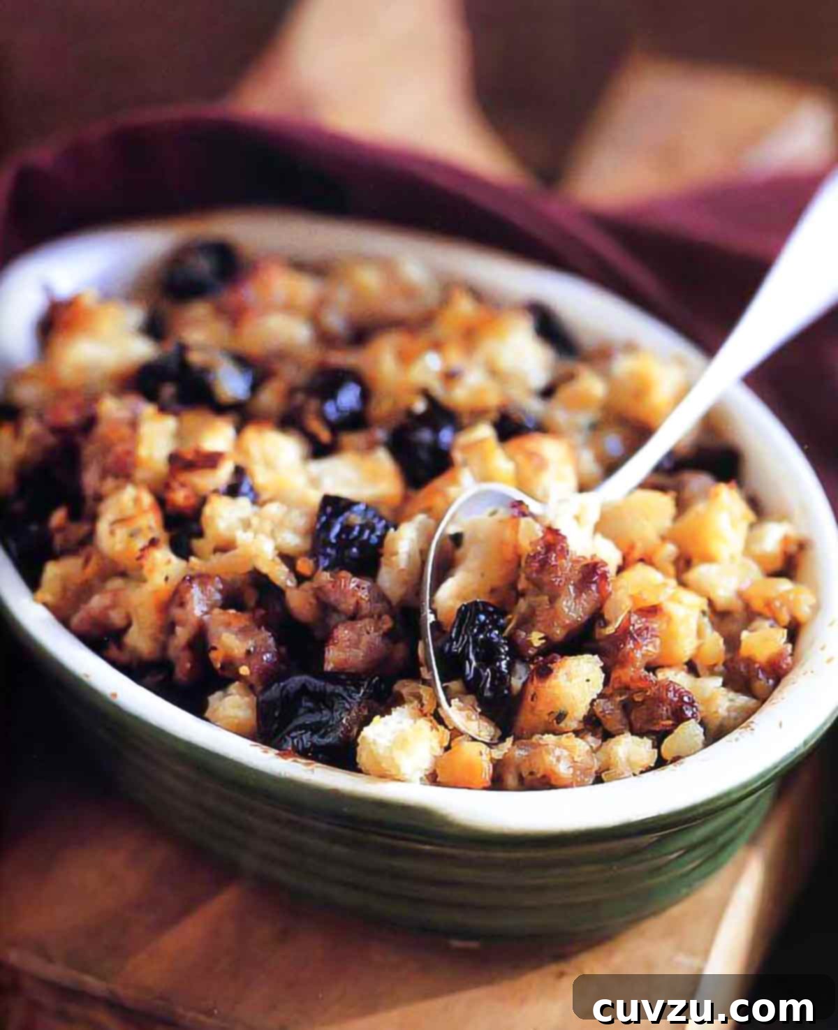 Sourdough dressing with sausage and prunes in a green ceramic dish with a large serving spoon.