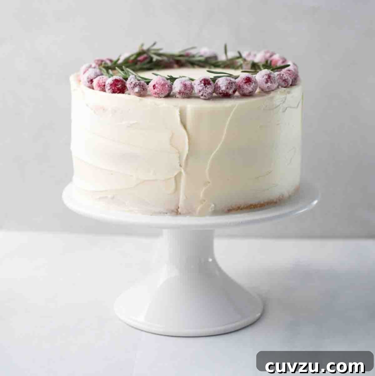 A whole frosted white Christmas cake topped with sugared cranberries and rosemary sprigs on a white cake stand.