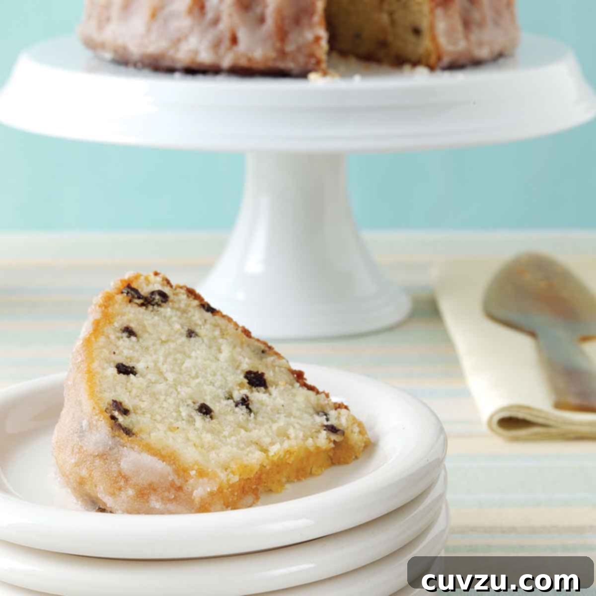 A plate with a slice of pound cake with currants, and a cake stand in the background with the remaining cake.