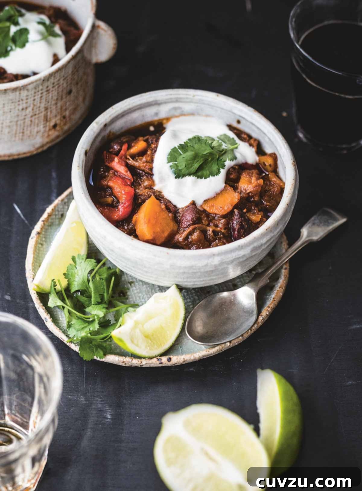 A bowl of shredded beef chili with sweet potatoes on a plate containing a spoon, two lime wedges, and cilantro leaves.