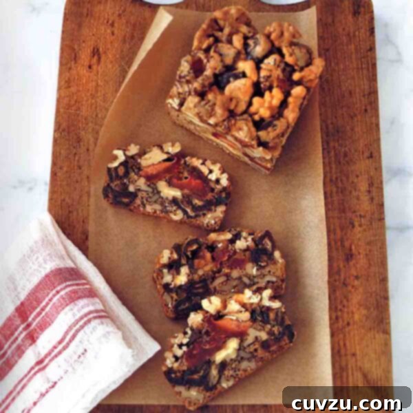 Dried fruit and nut cake in slices on a wooden cutting board with parchment paper, beside a red and white dish towel.
