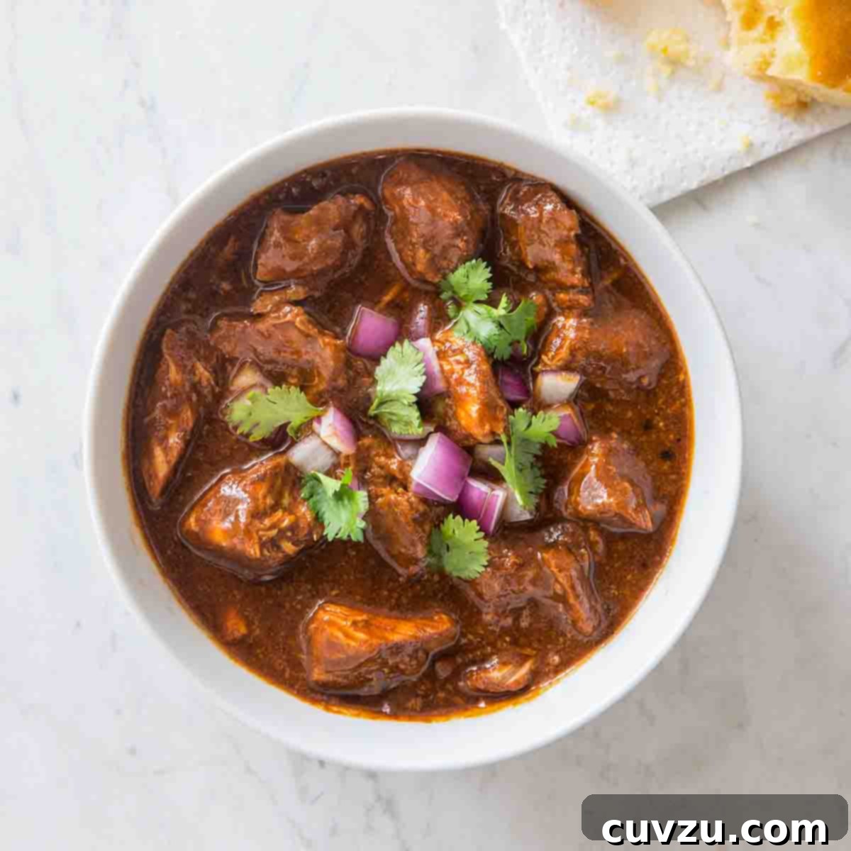 A white bowl filled with New Mexican red pork chili, garnished with red onion and cilantro.