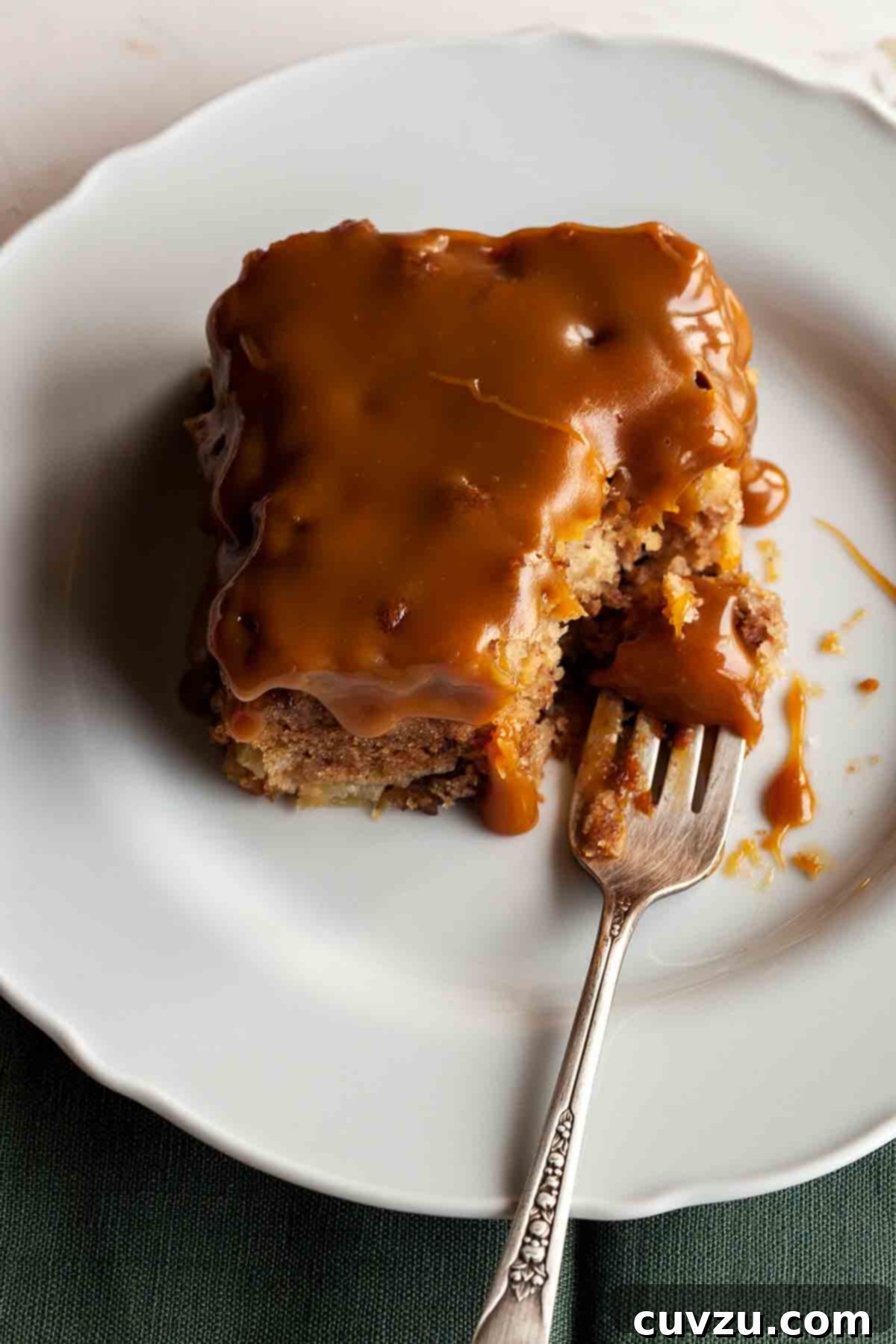A square of Edna Lewis's apple cake with caramel glaze on a white plate with a fork, highlighting its moist texture and rich caramel topping.