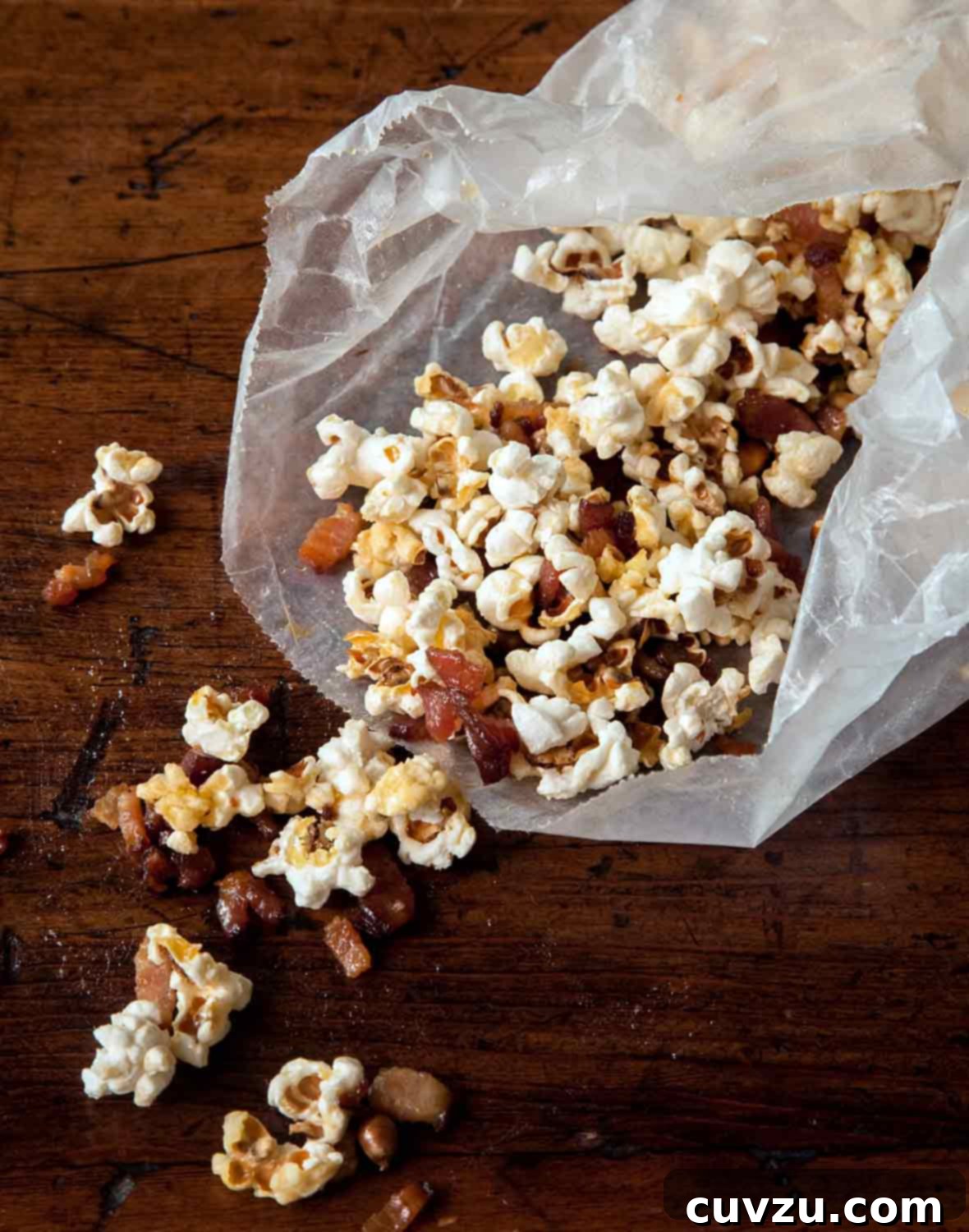 An open wax paper bag overflowing with fluffy popcorn, adorned with golden bacon bits and a drizzle of maple syrup, artfully spilled onto a surface.