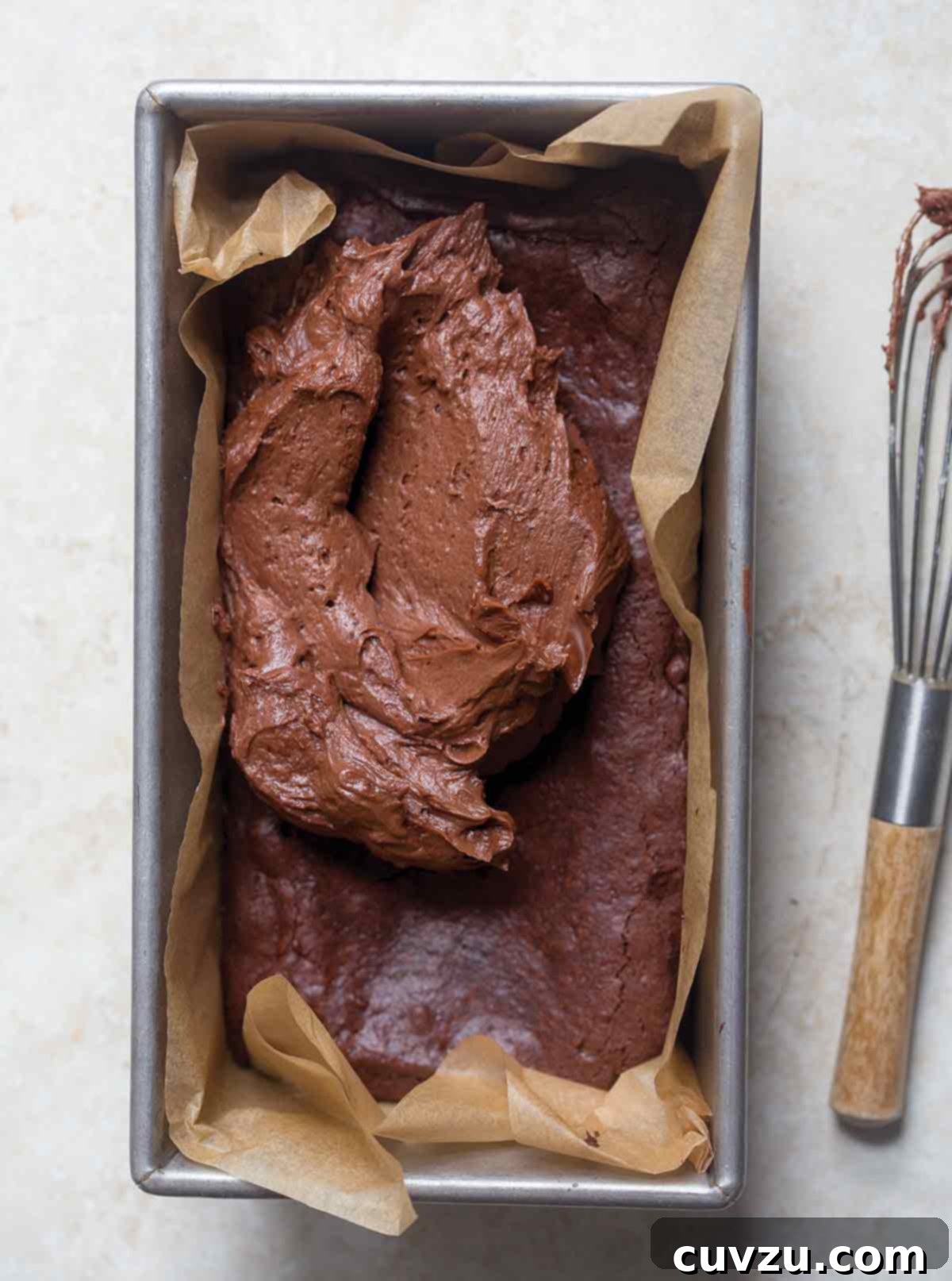A parchment lined loaf pan filled with partially frosted small batch brownies and a wire whisk on the side.