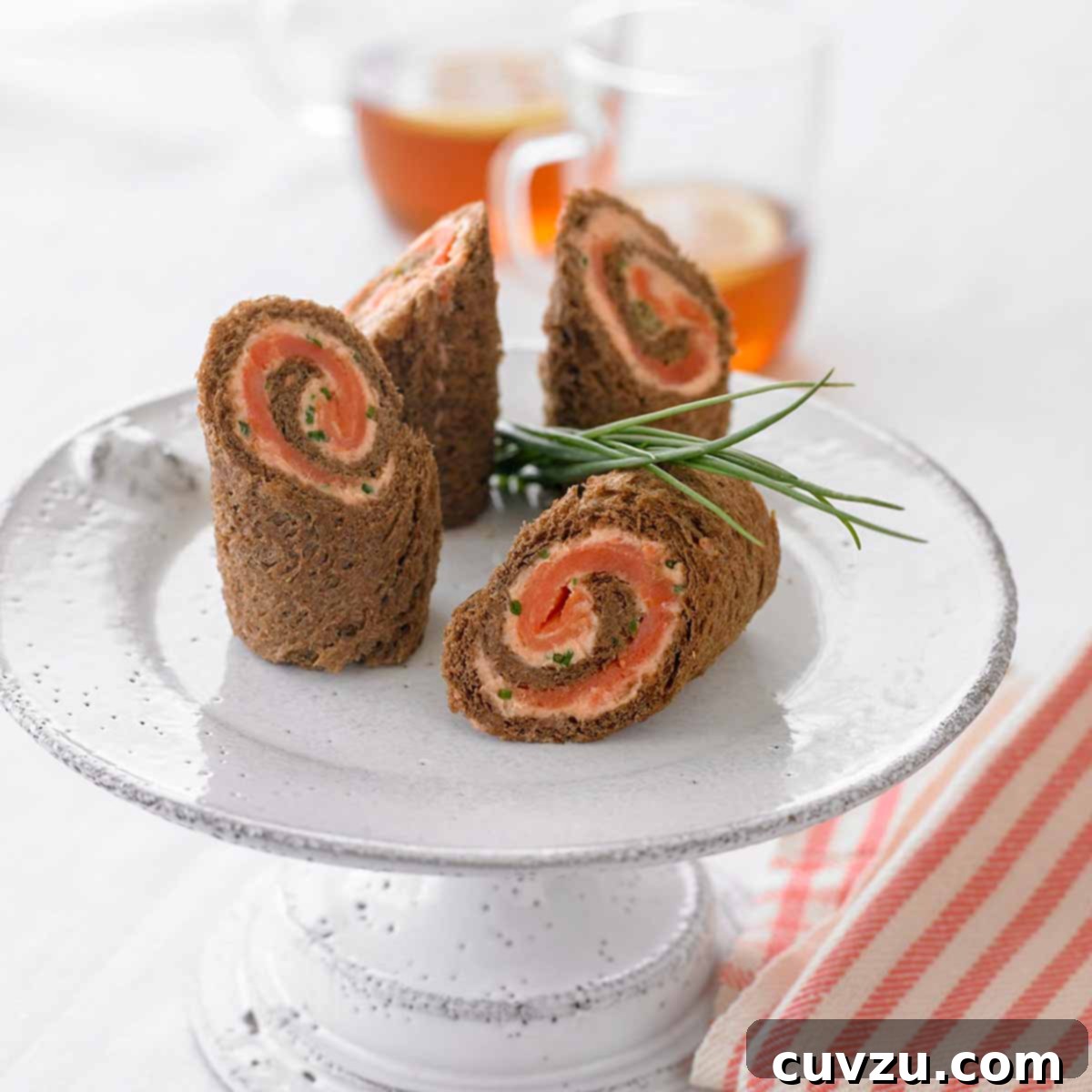 A white cake stand with four spirals of pumpernickel bread, salmon mousse, and smoked salmon, arranged beautifully.