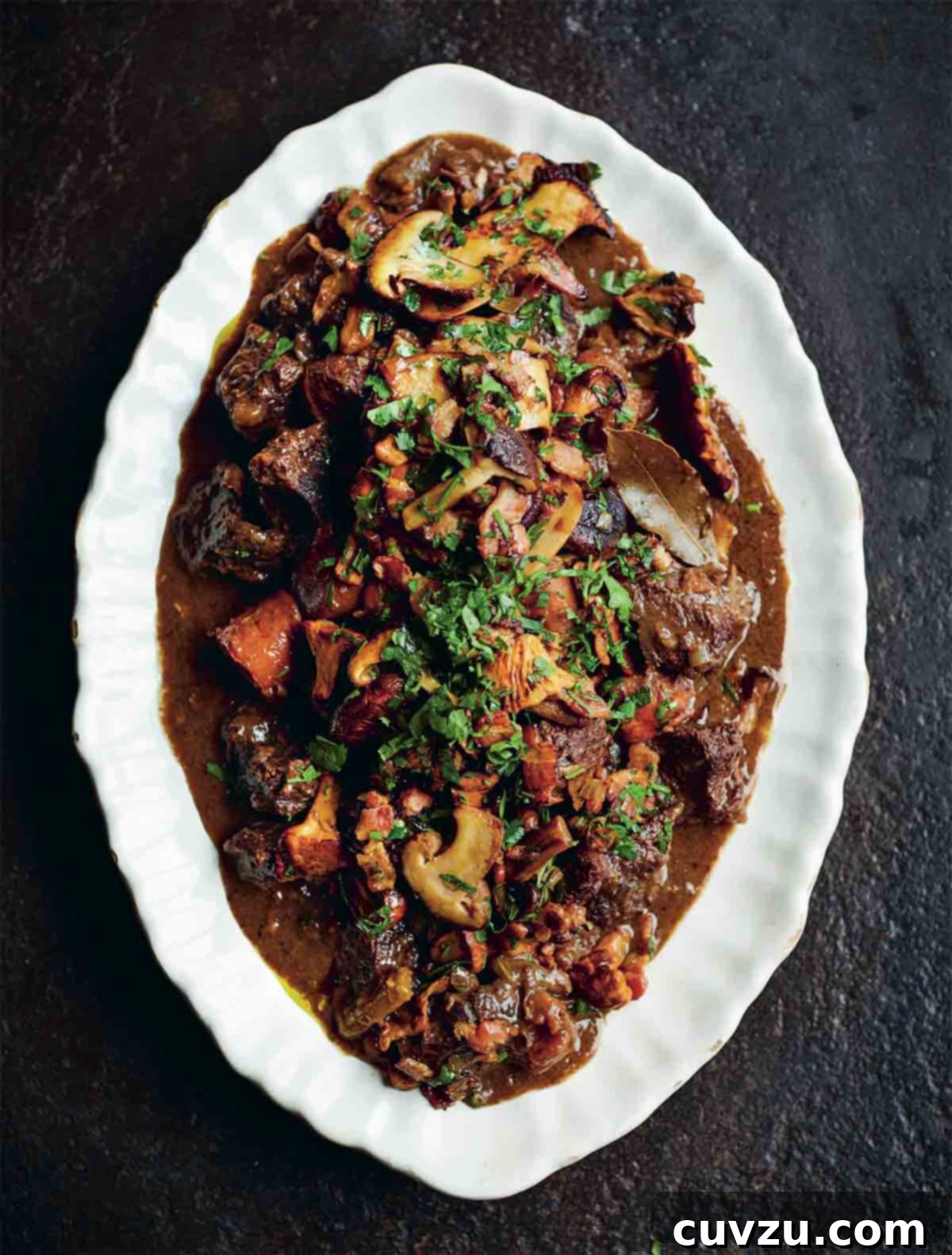A white platter with Jamie Oliver's Beef Bourguignon, filled with tender beef, carrots, and mushrooms, garnished with herbs.