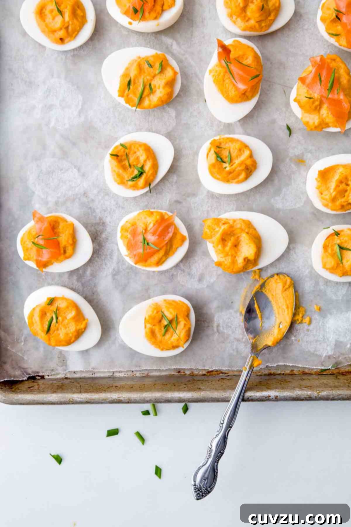 Sixteen Sriracha deviled egg halves and a teaspoon on a parchment-lined baking sheet, garnished with herbs.